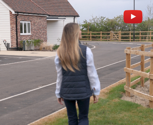 Woman walking away from the camera along a suburban street with a brick house and wooden fence; a YouTube play button badge is in the top-right corner.