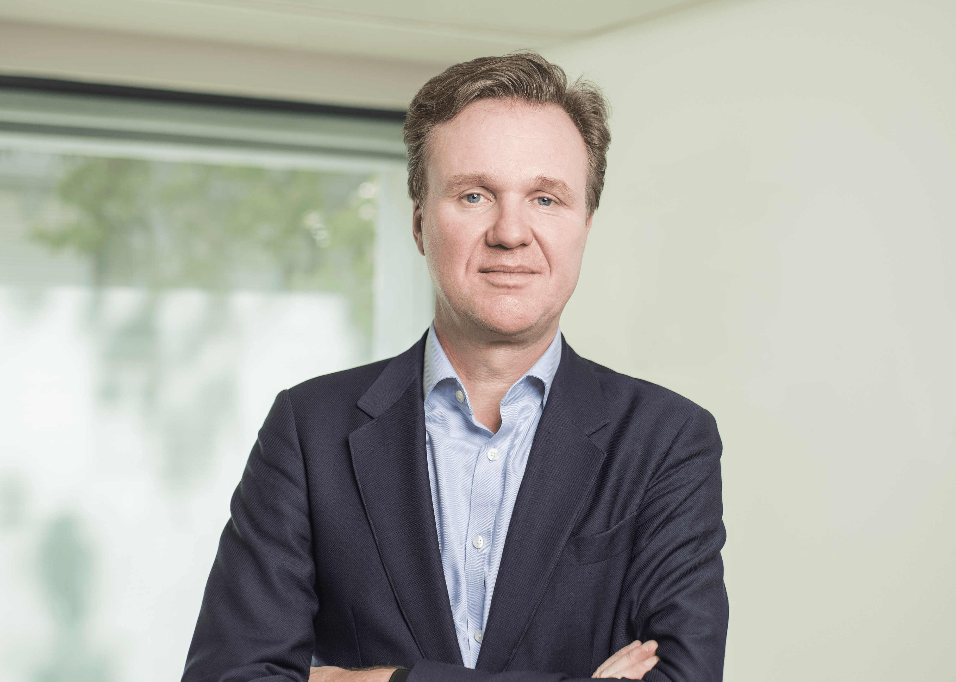 Portrait of a man in a navy blazer and light blue shirt with arms crossed in a bright office setting
